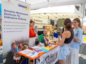 Das Foto zeigt den Stand des AW-ZIB und der Fachschaft Sonderpädagogik bei den PH Days. Im Vordergrund ist ein langer Tisch, auf dem sich verschiedeneDinge befinden: das Glücksrad, bunte Karten, auf denen die Fragen geschrieben stehen, Preise. Vor dem Tisch stehen zwei Studentinnen, hinter dem Tisch sind zwei Personen, davon sitzt eine Person im Rollstuhl. Links neben dem Stand ist ein großer Banner. Auf disem steht: Annelie-Wellensiek-Zentrum für Inklusive Bildung.