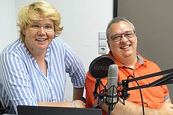 Das Foto zeigt zwei Personen nebeneinander sitzen. Beider lächeln in die Kamera und vor ihnen ist ein Mikrofon befestig.