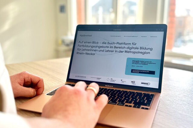 Der Fokus liegt auf einem Laptop, auf dem die Website digi@school zu sehen ist. Links im Bildrand ist eine Person unscharf zu sehen, die den Laptop bedient.