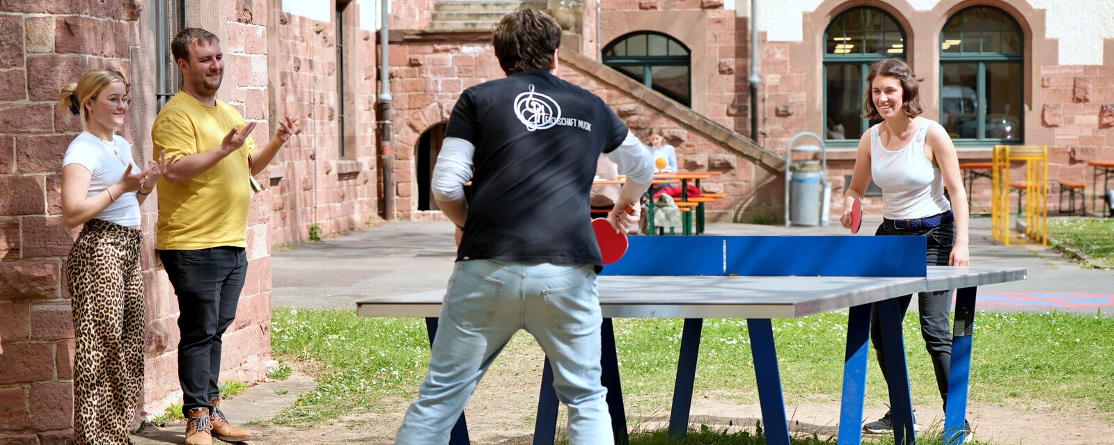 Zu sehn sind zwei Menschen beim Tischtennis Spielen. Ein Mann trägt ein T-Shirt mit der Aufschrift "Fachschaft Musik".