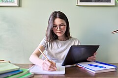 Das Bild zeigt eine junge Frau am Schreibtisch. Sie hält ein Tablet in der einen Hand und notiert mit der anderen etwas auf einen Block.