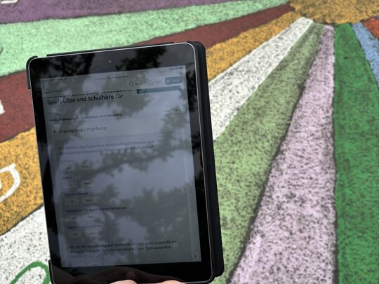 Das Foto zeigt ein Tablet mit dem Fragebogen der Wissenschaftler, im Hintergrund ist ein bemalter Fußboden eines Schulhofs