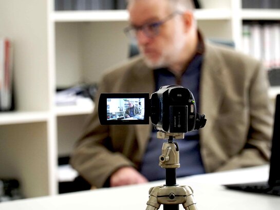 Das Bild zeigt Hartmut Kabelitz an seinem Arbeitsplatz. Vor Herr Kabelitz steht eine Videokamera.