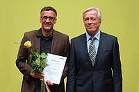 Michael Neuberger und Karl-Heinz Dammer. Neuberger hat die Lehrpreis-Urkunde und eine gelbe Rose in der Hand. Beide schauen freundlich in die Kamera.