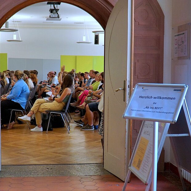 Blick vom Foyer in die vollbesetzte Aula. Davor ein Schild, der die Teilnehmer:innen zu der Veranstaltung Ab ins Ref begrüßt.