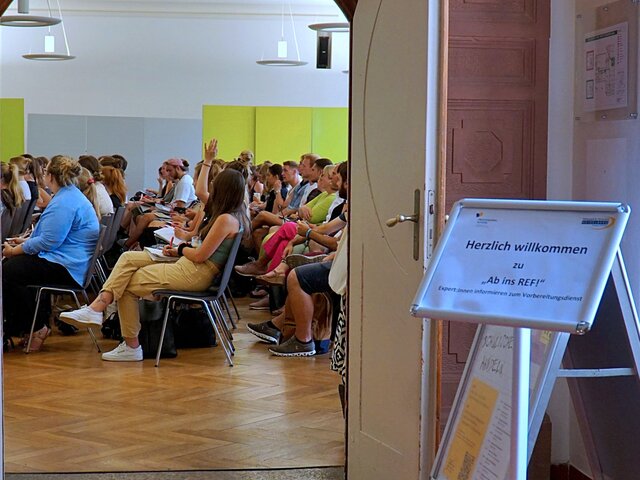 Blick vom Foyer in die vollbesetzte Aula. Davor ein Schild, der die Teilnehmer:innen zu der Veranstaltung Ab ins Ref begrüßt.