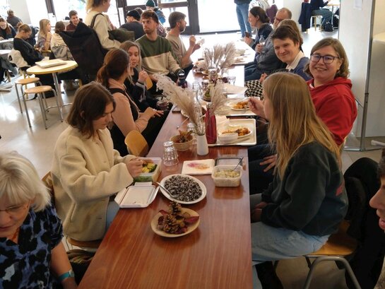 In der Neubaucafeteria sitzen Mitarbeitende und Stdudierende gemeinsam an einer langen Tafel. Auf dem Tisch sind Dekoration und vegane Snacks platziert und