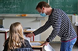 Das Symbolbild zeigt eine Schülerin an ihrem Arbeitsplatz in der Schule. Daneben steht ein Lehrer mit einer Mund-Nasen-Bedeckung und gibt ihr Arbeitsblätter. Im Hintergrund ist eine beschriebene Kreidetafel. Copyright Pädagogische Hochschule Heidelberg