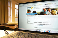 Laptop mit Startseite der AW-ZIB-Website in einfacher Sprache. Links neben dem Laptop liegt ein Block mit Stift. Im Hintergrund sieht man unscharf einen Hörsaal.
