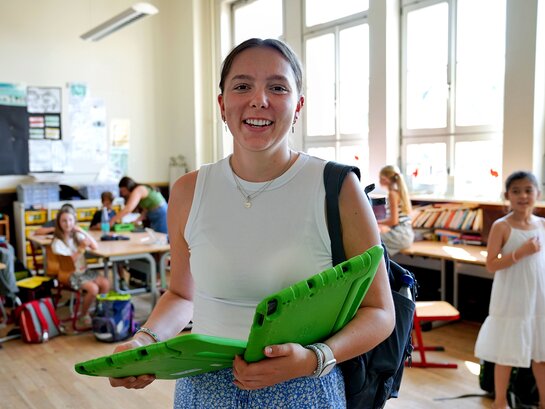 Das Foto zeigt eine Lehrkraft mit zwei Tabletts in der Hand. Im Hintergrund sind Schüler:innen.