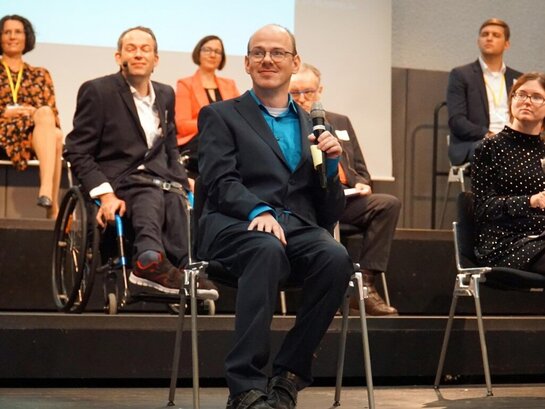 Das Foto zeigt verschiedene Menschen auf einer Bühne mit einem Mikrofon in der Hand.