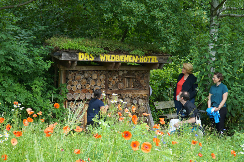 Das Bild wurde im Ökogarten aufgenommen. Man sieht ein Wildbienen-Hotel. Davor sitzt links eine junge Frau; man sieht sieh von hinten. Rechts im Bild stehen zwei Frau; neben ihnen sitzt ein Mann im Rollstuhl. Man sieht zudem im Hintergrund viele grüne Bäume. Im Vordergrund ist eine wilde, hochgewachsene Blumenwiese zu sehen. 