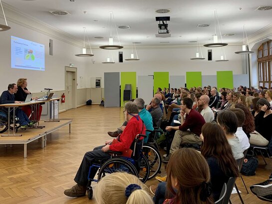 Das Bild zeigt Thorsten Lihl und Karin Terfloth, die in der Aula der Pädagogischen Hochschule Heidelberg einen Vortrag vor vielen Menschen halten.