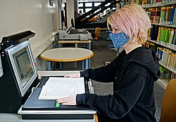 Das Bild zeigt eine Studentin am Laptop mit Maske in der Bibliothek.