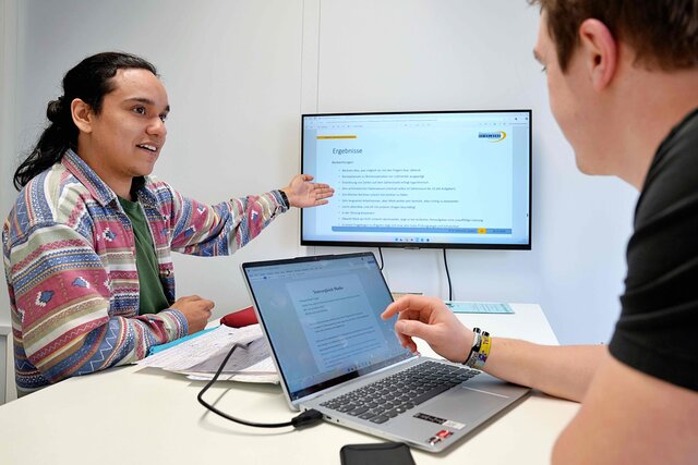 Ein junger Mann erklärt einem Studenten Inhalte zu wissenschaftlichen und zentrale Einrichtungen, die Themen wie Inklusion, Digitalisierung und Nachhaltigkeit.