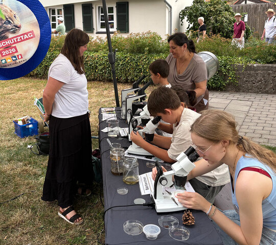 Kider und Erwachsenen sitzen an einem Tisch und arbeiten mit Mikroskopen. Eine Studierende erklärt dazu.