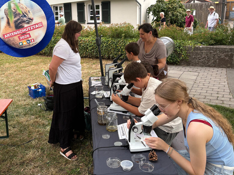 Kider und Erwachsenen sitzen an einem Tisch und arbeiten mit Mikroskopen. Eine Studierende erklärt dazu.
