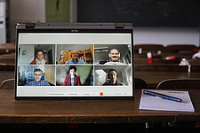 Das Bild zeigt einen Seminarraum mit einem Tablet. Au dem Tablet sind die Teilnehmer der Online-Veranstaltung