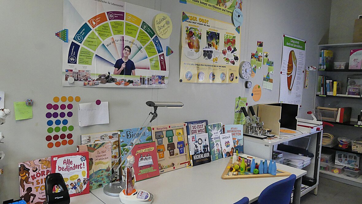 Zu sehen ist ein Bereich der Didaktischen Werkstatt der Alltagskultur und Gesundheit: viele Kinderbücher stehen auf einem Schreibtisch, Poster hängen an der Wand, 