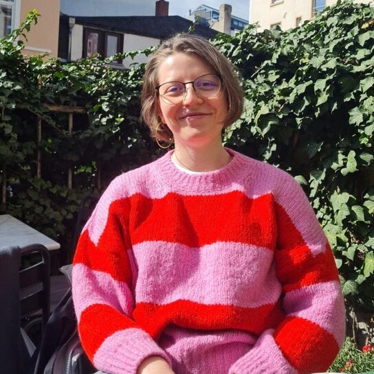 Clara sitzt im Grünen und lächelt in die Kamera. Sie hat einen pink-rot-gestreiften Pulli an.