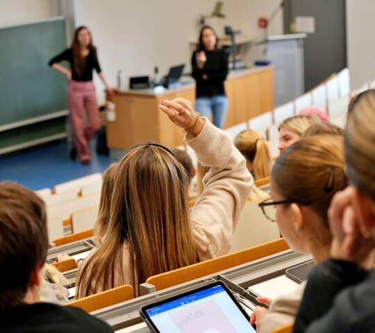 Studierende sitzen im Hörsaal, vorne steht die Dozentin.
