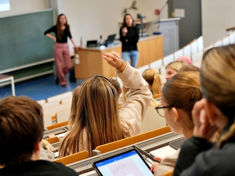 Studierende sitzen im Hörsaal, vorne steht die Dozentin.