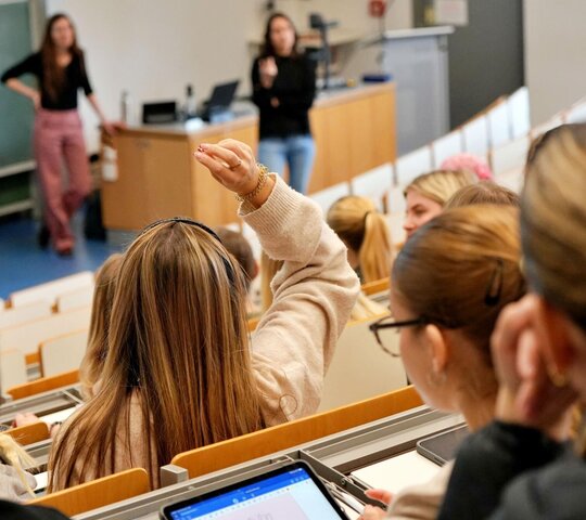 Studierende sitzen im Hörsaal, vorne steht die Dozentin.