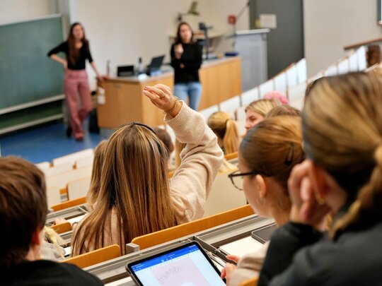 Studierende sitzen im Hörsaal, vorne steht die Dozentin.