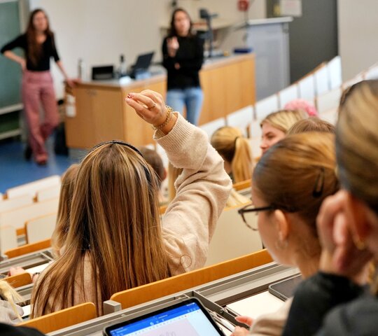 Studierende sitzen im Hörsaal, vorne steht die Dozentin.