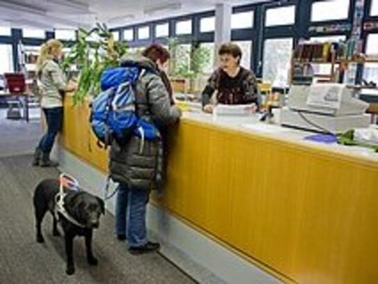 Eine Frau steht an einem Tresen und spricht mit einem Mann. Die Frau hat einen Assistenzhund an der Leine dabei.