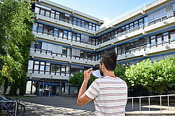 Das Symbolbild zeigt einen jungen Mann, der sich gerade eine Atemschutzmaske auszieht. Er steht vor dem Neubau der Hochschule und ist von hinten zu sehen. Copyright Pädagogische Hochschule Heidelberg.