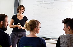 Das Bild zeigt eine Frau die an einer digitalen Tafel steht und drei weitere Personen, die sitzen und zuhören. Copyright Pädagogische Hochschule Heidelberg
