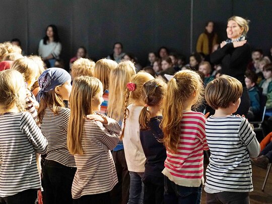 Eine Gruppe von stehenden Kindern, alle in gestreiften Pullis, von hinten. Vor ihnen steht - unscharf - eine junge Lehrerin, die Grundschullehramt an der PHHD studiert hat. 