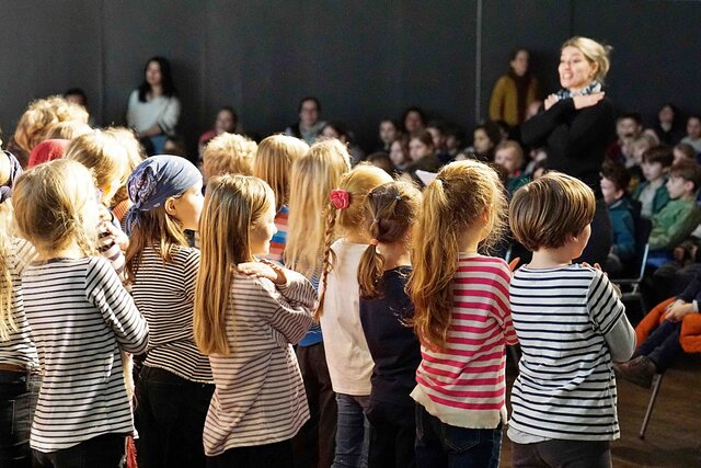 Eine Gruppe von stehenden Kindern, alle in gestreiften Pullis, von hinten. Vor ihnen steht - unscharf - eine junge Lehrerin, die Grundschullehramt an der PHHD studiert hat. 