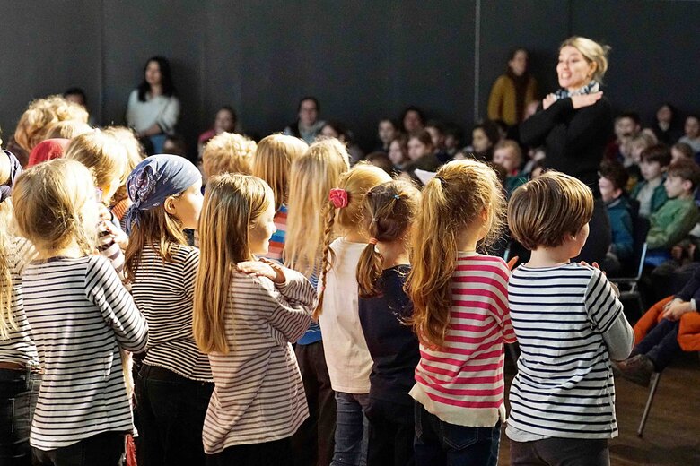 Eine Gruppe von stehenden Kindern, alle in gestreiften Pullis, von hinten. Vor ihnen steht - unscharf - eine junge Lehrerin, die Grundschullehramt an der PHHD studiert hat. 