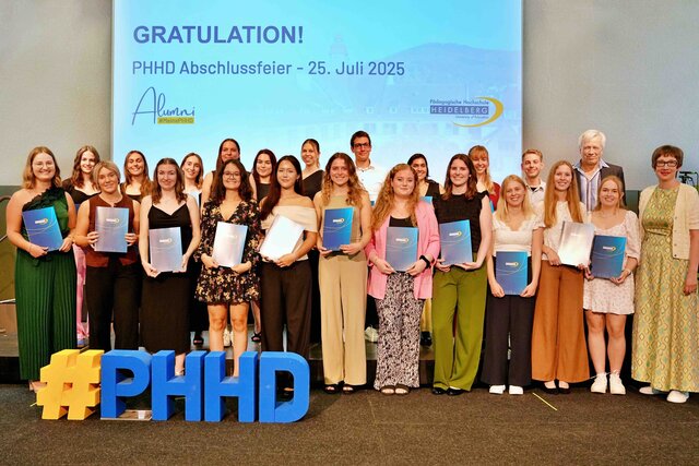 Zu sehen sind Alumni, die auf einer Bühne stehen und ihre Bestehensbriefe in PHHD-Mappen hochhalten. Gemeinsam mit Rektorin Vach & Prorektor Dammer (a.D.), welche ihnen die Mappen überreicht haben.