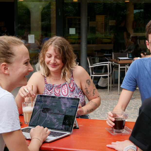 Gutgelaunte Studierende sitzen mit Laptops und Getränken draußen an einem Biertisch