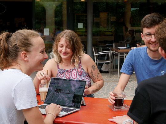 Gutgelaunte Studierende sitzen mit Laptops und Getränken draußen an einem Biertisch