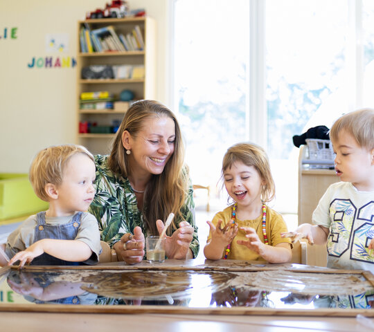 Eine Erzieherin sitzt mit drei Kindern an einem Tisch und spielt und arbeitet mit ihnen.