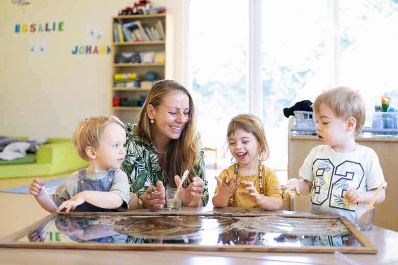 Eine Erzieherin sitzt mit drei Kindern an einem Tisch und spielt und arbeitet mit ihnen.