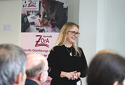Auf dem Foto sieht man eine lachende Frau vor dem "Zukunfts-Orientierungs-Akademie" (ZOrA)- Plakat.