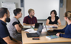 Das Foto zeigt eine Gruppe Studierender die sich an Tischen mit Laptops gegenüber sitzt.