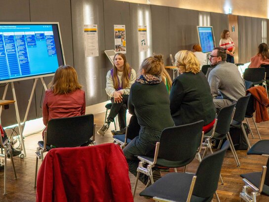 Das Bild zeigt einen Austausch während des Tags der Vielfalt in der Mehrzweckhalle der Pädagogischen Hochschule Heidelberg. Mehrere Personen sitzen im Halbkreis vor einem großen Monitor.