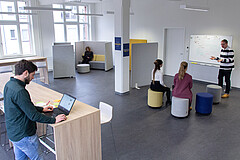 Das Symbolbild zeigt den Design Thinking Raum der Hochschule. Es stehen mehrere Personen an verschiedenen stellen und arbeiten am Laptop bzw. unterhalten sich.