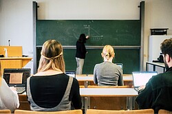 Das Foto zeigt Studierende in einem Seminarraum. Im Hintergrund ist erkennbar, dass der Dozent etwas an die Tafel schreibt.