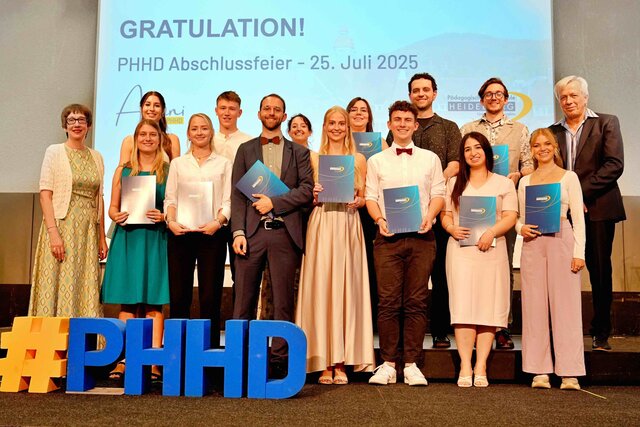 Zu sehen sind Alumni, die auf einer Bühne stehen und ihre Bestehensbriefe in PHHD-Mappen hochhalten. Gemeinsam mit Rektorin Vach & Prorektor Dammer (a.D.), welche ihnen die Mappen überreicht haben.