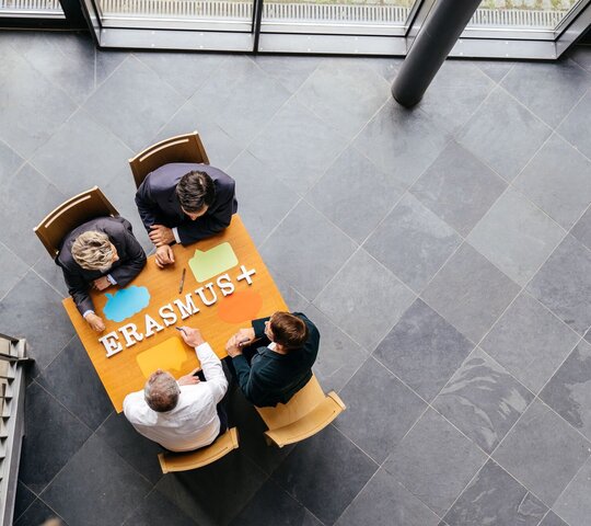 Aus der Vogelperspektive sehen wir vier Personen an einem Informationstisch zu Angeboten von Erasmus+ sitzen.