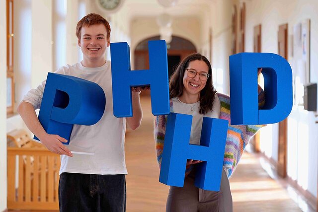 Eins Student und eine Studentin der Pädagogischen Hochschule Heidelberg halten die Buchstaben PHHD hoch. Sie befinden sich im Altbau.