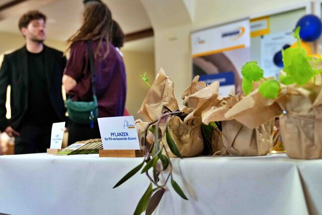 Zu sehen ist ein Tisch mit Pflanzen. verschwommen im Hintergrund eine Alumna, die sich eine Pflanze mitnimmt und mit einem weiteren Gast unterhält.