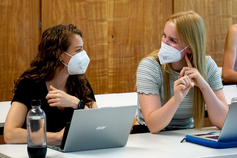 Das Symbolbild zeigt zwei Studentinnen. Sie sitzen in einem Seminarraum der Hochschule. Vor ihnen steht jeweils ein Laptop. Sie schauen sich freundlich an. Beide tragen corona-bedingt einen Mund-Nase-Schutz.